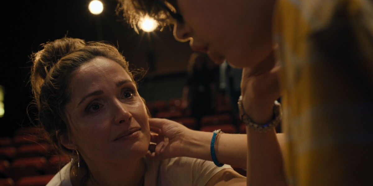 A person with a concerned expression looks up at another person who is gently touching their face, with theater seats and blurred lights in the background.