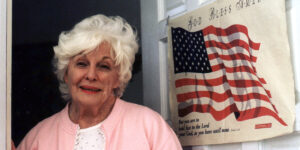 Arthur Dong’s LGBTQ Stories Blu-Ray Review – Essential Pride Month Viewing An elderly woman with white hair stands beside an American flag and a "God Bless America" poster, wearing a pink cardigan over a white blouse.