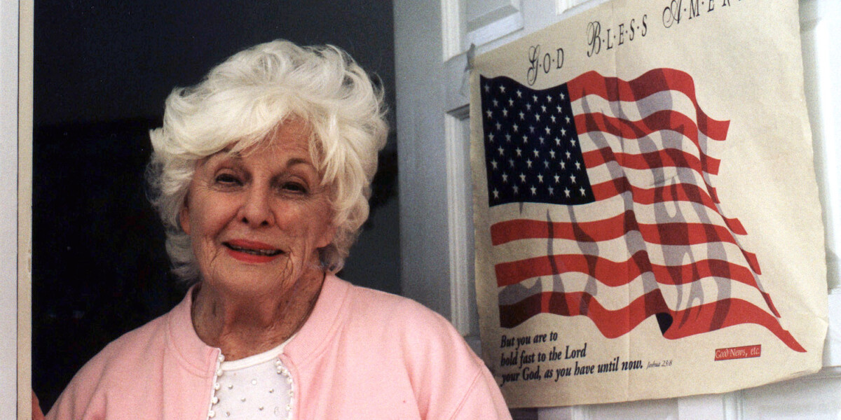 An elderly woman with white hair stands beside an American flag and a "God Bless America" poster, wearing a pink cardigan over a white blouse.