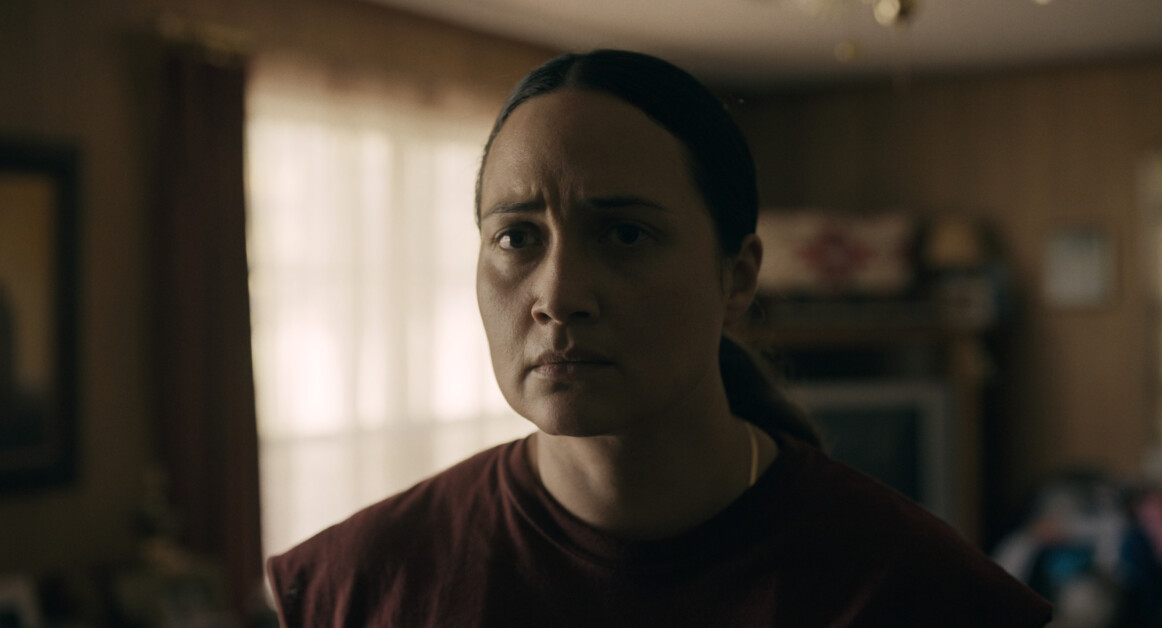 A person with a serious expression stands in a dimly lit room with beige walls, a window with sheer curtains, and an old television in the background.