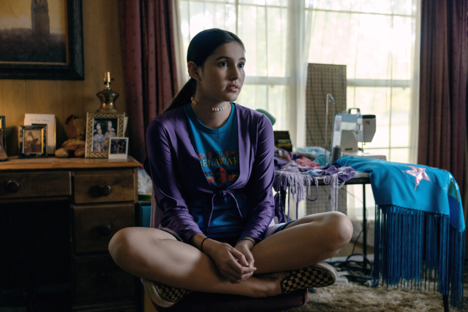 A young person sits cross-legged on a chair in a cluttered room with a sewing machine and fabric.