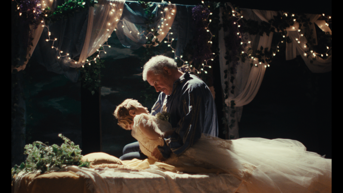 An older man cradles a younger woman in a white dress on a decorated bed with string lights and draped fabric in the background.