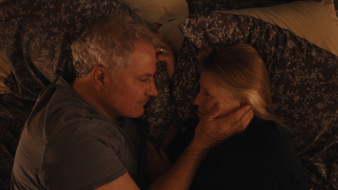 Two people lie in bed facing each other, with the person on the left gently cradling the face of the person on the right. Both are resting on patterned pillows.