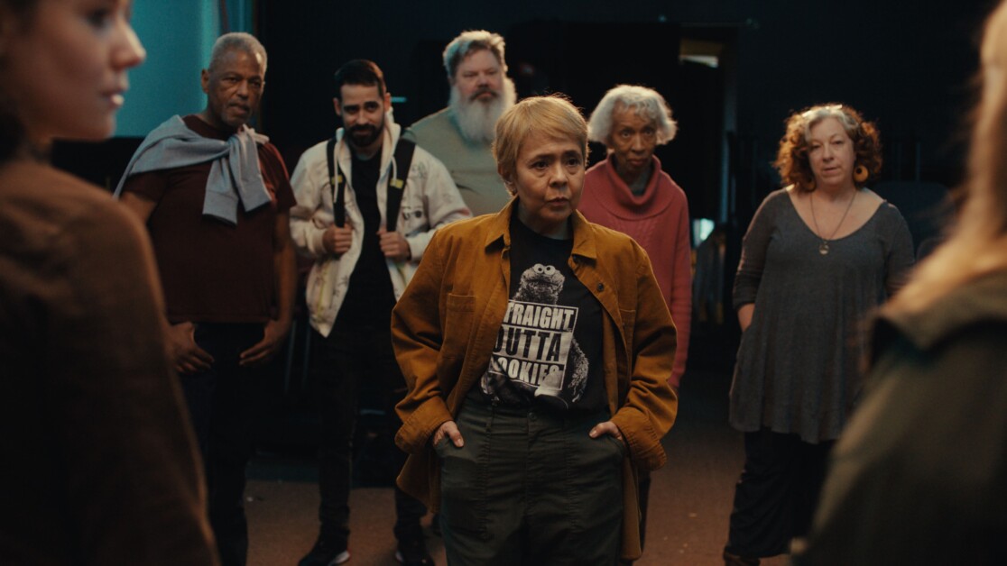 A group of people stand in a semi-circle in a dimly lit room. One person in the center, wearing a brown jacket and printed t-shirt, is speaking or leading the group.