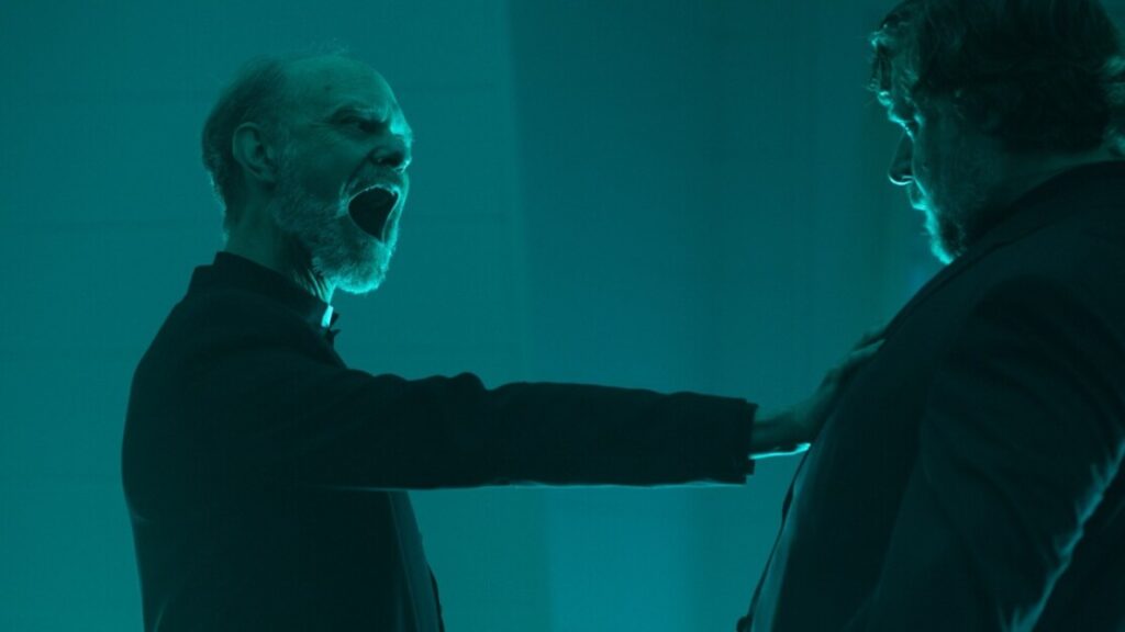 An elderly man with an open mouth holds his hand on another man's chest in a dimly lit greenish-blue room.