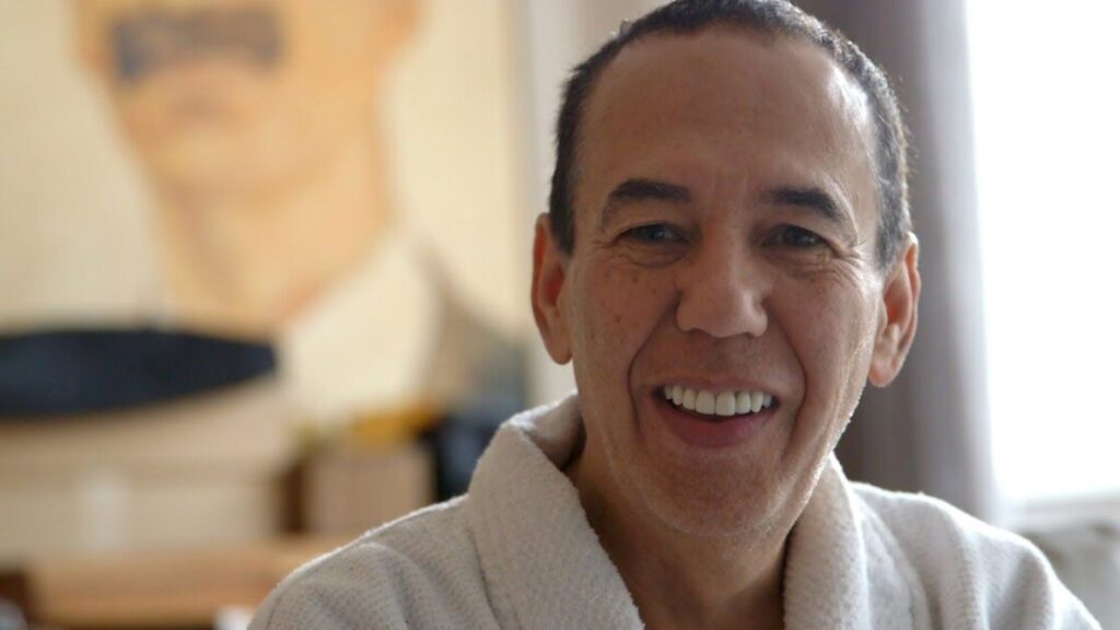 A man wearing a white bathrobe smiles at the camera with a blurred portrait of another man in the background.