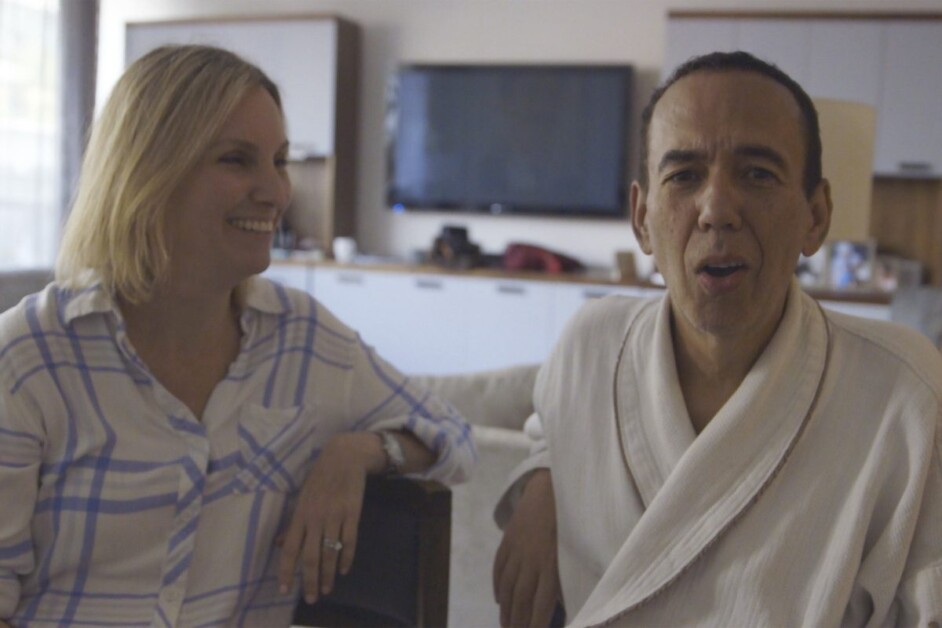 A woman in a plaid shirt smiles at a man in a bathrobe who is speaking. They are in a room with a TV and cabinets in the background.