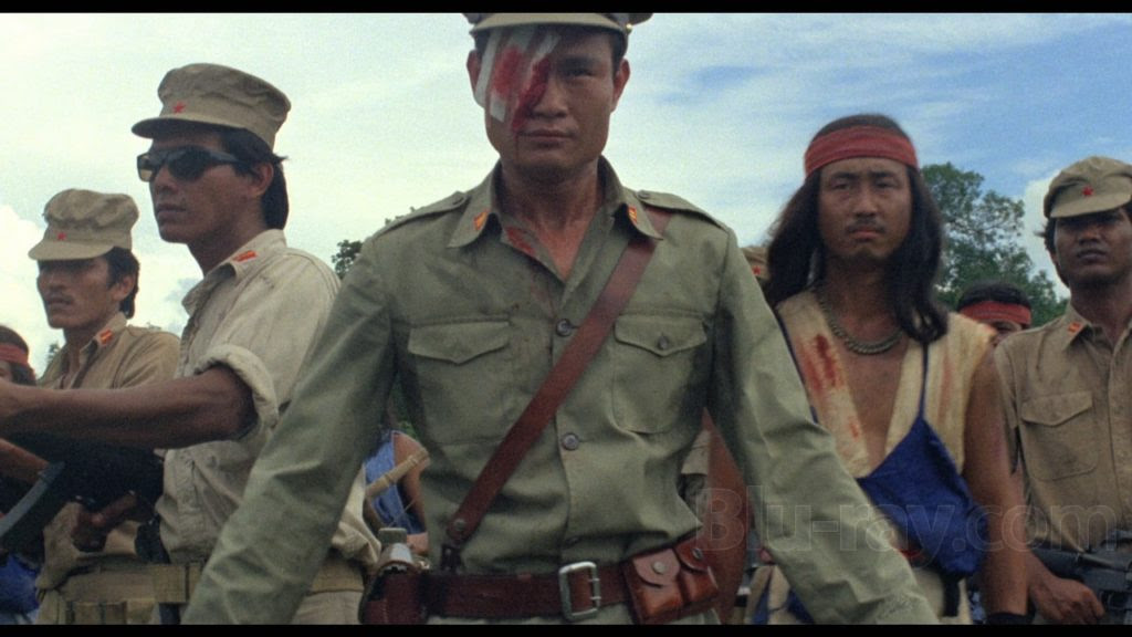 A group of people in military attire, with one man at the center wearing an eye patch and a bloodstained uniform. They appear serious and are outdoors.