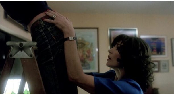 A person wearing a blue shirt steadies someone else who is standing on a ladder in a room with framed pictures on the wall.