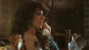 A woman with curly hair is speaking on a corded telephone, looking to her right. She wears a dark top and is indoors with a blurred background.