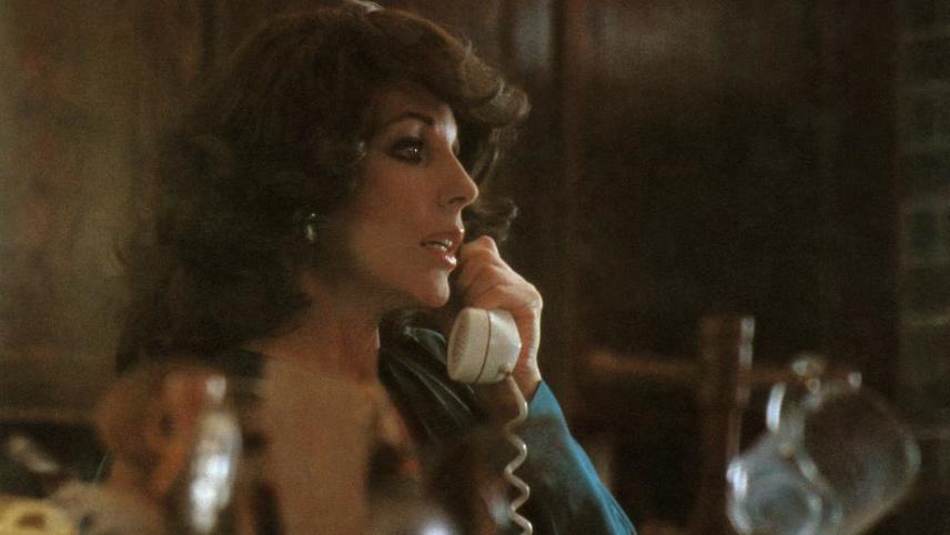 A woman with curly hair is speaking on a corded telephone, looking to her right. She wears a dark top and is indoors with a blurred background.