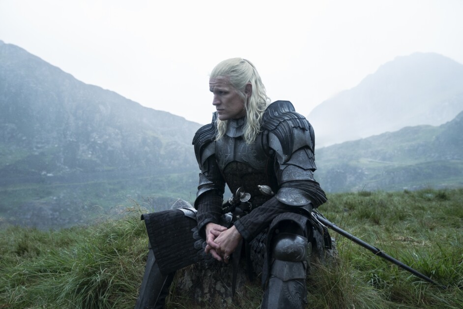 A person with long blonde hair, wearing dark armor, sits on a rock in a misty mountainous landscape, looking contemplative.