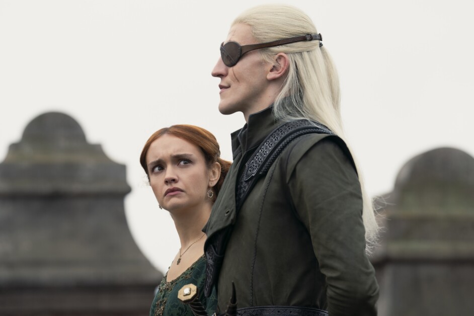 Two characters are standing outdoors, one with long blonde hair and an eye patch, the other with auburn hair, appearing concerned. Stone structures are visible in the background.
