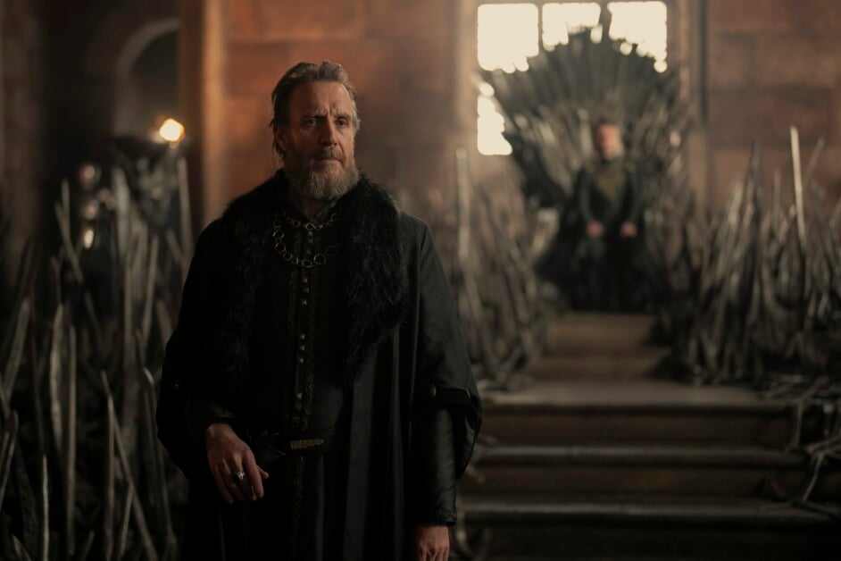 A man in medieval attire stands in a dimly lit room with a large throne made of swords in the background.