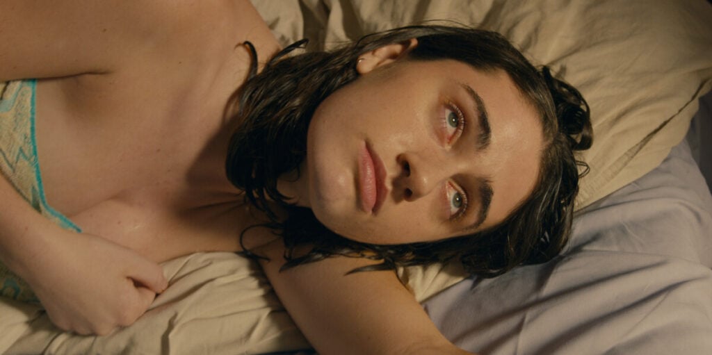 A woman with wet hair lies on a bed, looking contemplative. She is covered with a beige sheet and her head rests on a pillow.