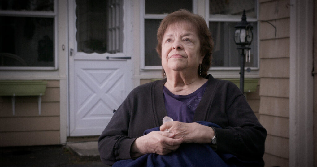 An older woman with short brown hair sits outside on a porch. She is wearing a dark sweater and holding a blanket, looking off into the distance. The background shows a house with a white door.