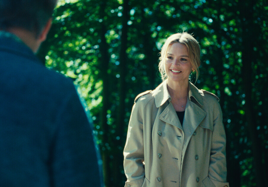 A woman in a beige trench coat smiles while talking to a man in a blue jacket. They are outdoors with trees in the background.