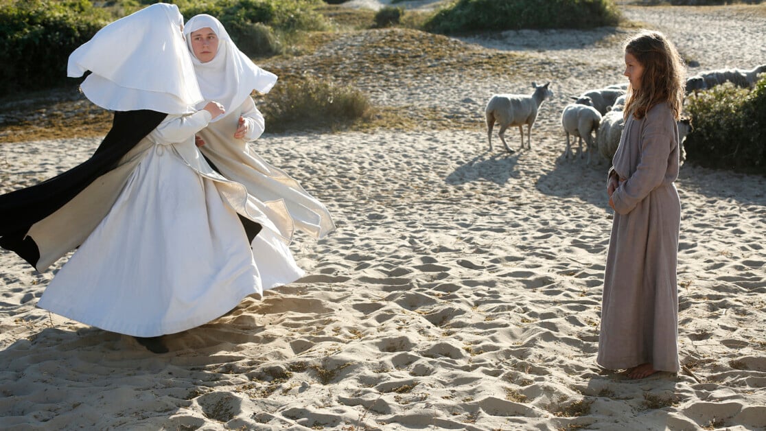 A woman dressed in a white nun's habit and a girl in a simple tunic stand on a sandy area with sheep grazing in the background.