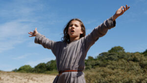 ‘Jeannette: The Childhood Of Joan Of Arc’ Blu-Ray Review – Rocking Out For The Lord A child with long hair in a gray tunic stands outdoors with arms outstretched, appearing to shout or sing. Trees and a blue sky are in the background.