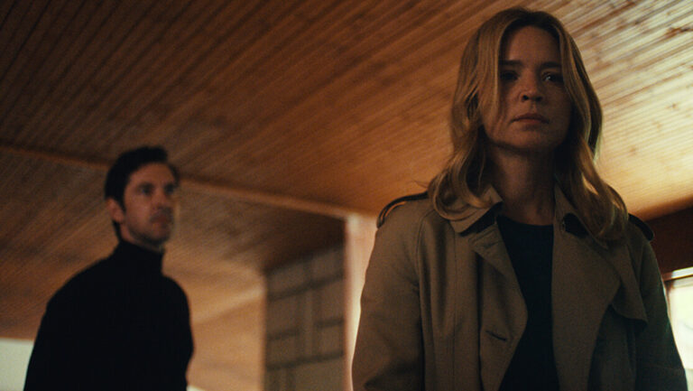 ‘Just The Two Of Us’ Review – Love Is A Forest With Too Many Trees A woman with blonde hair in a beige coat stands in the foreground, while a man in a dark turtleneck stands in the background in a wooden-ceilinged room.