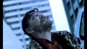 A man with wet hair and a beard looks up while standing between tall buildings, wearing a leopard print coat and a red shirt.