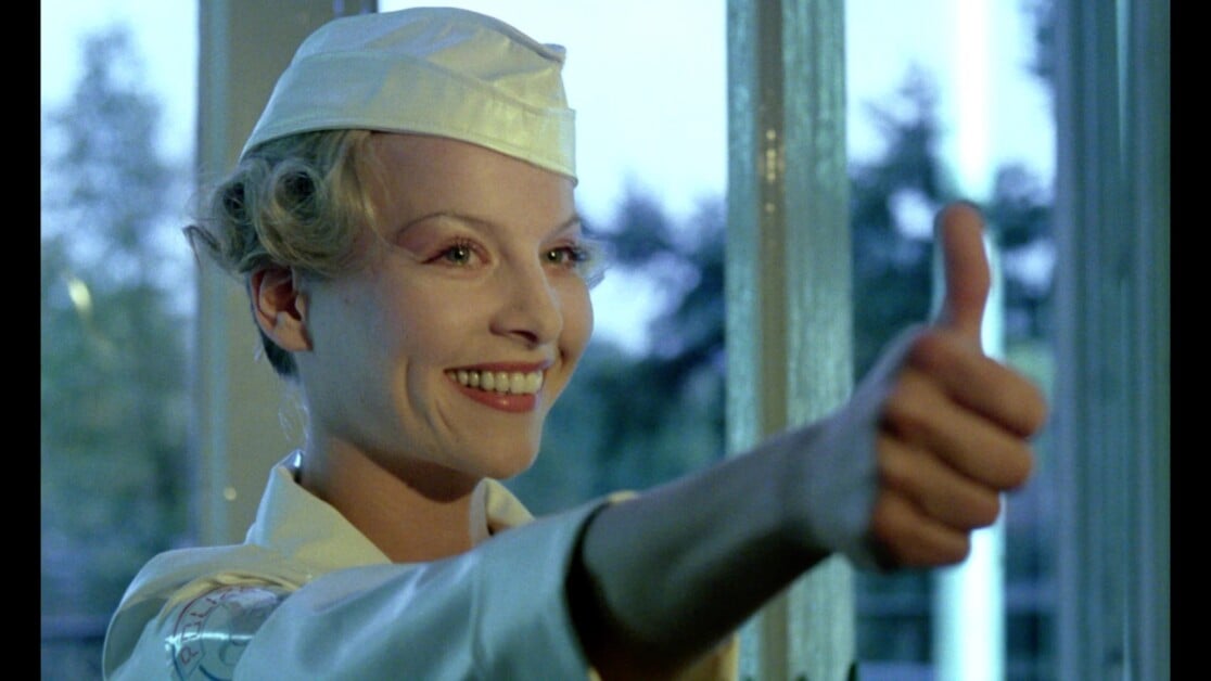 Person in a uniform hat smiles and gives a thumbs up gesture. Trees and window panes are visible in the background.