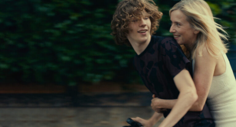 A person with curly hair rides a bike while another person holds on from behind. Both are smiling. They are outside with green foliage in the background.