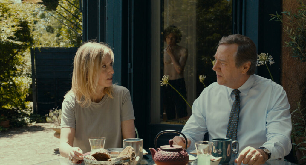 Two people sit at a table outside, engaged in conversation. Various food and drink items are on the table. In the background, a person is visible through a partially open door.