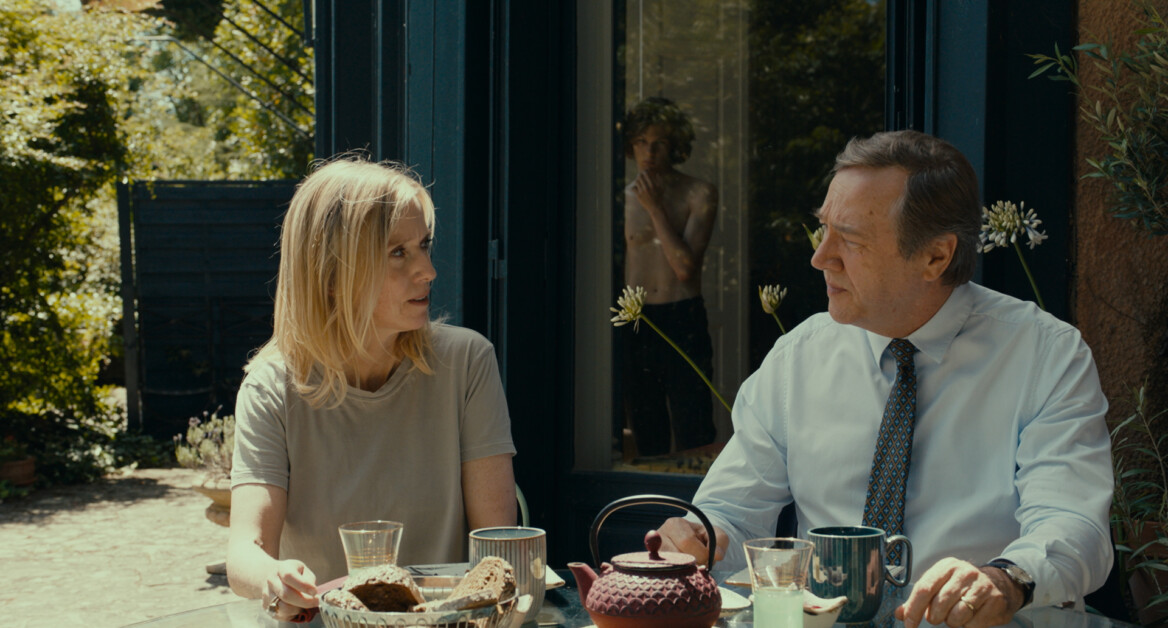 Two people sit at a table outside, engaged in conversation. Various food and drink items are on the table. In the background, a person is visible through a partially open door.