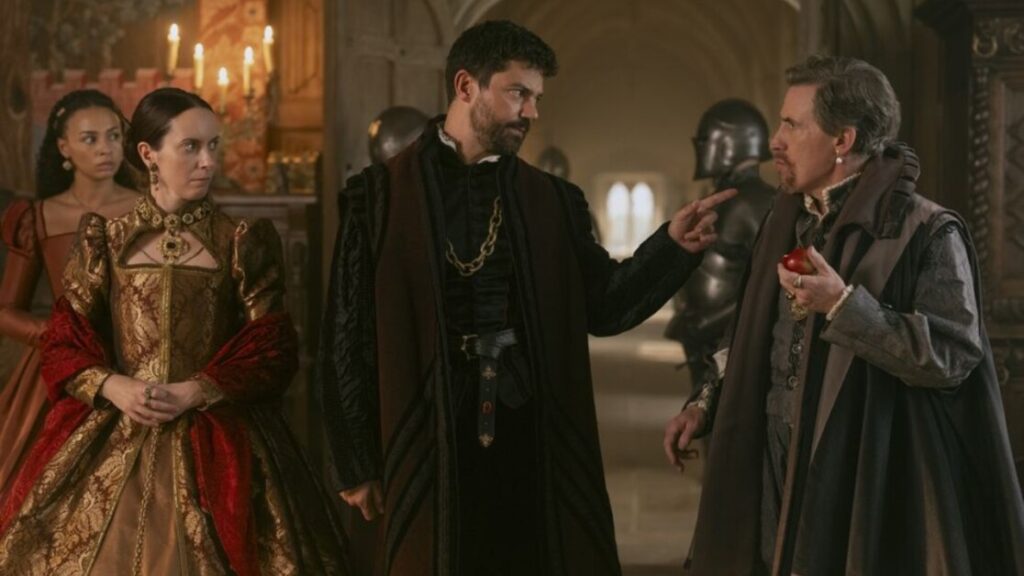 People in medieval costumes stand indoors; a bearded man points at another holding an apple, while two others observe. Armor is visible in the background.