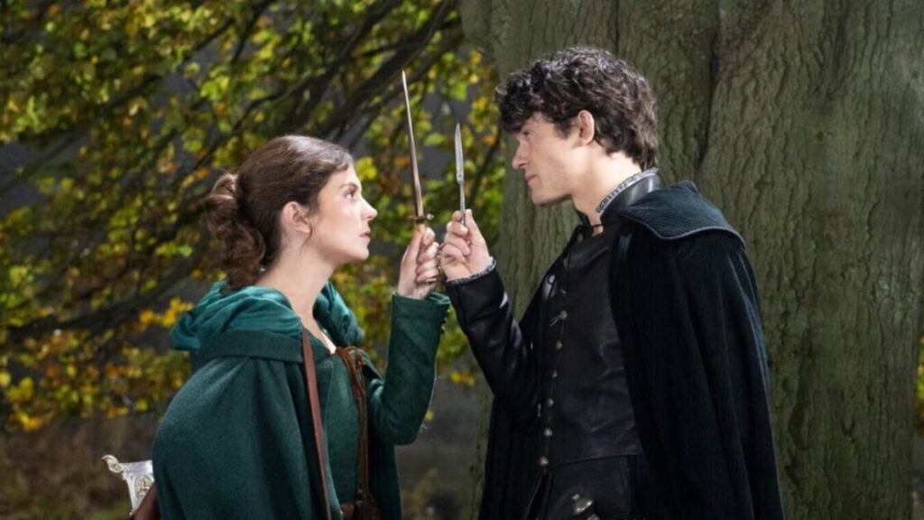 Two people dressed in medieval outfits face each other in a wooded area, holding small swords up in a seemingly intense conversation.