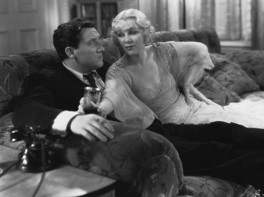 A man and woman in 1930s-style clothing sit on a patterned sofa; the man holds a glass, and the woman leans toward him while looking into his eyes.