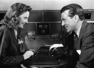A woman and a man are sitting across from each other at a table, smiling. The woman appears to be typing on a typewriter.