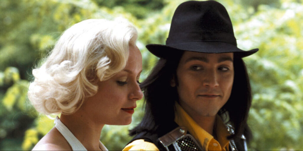 A woman with blonde hair and a man with long black hair and a hat standing outdoors, facing slightly toward each other.
