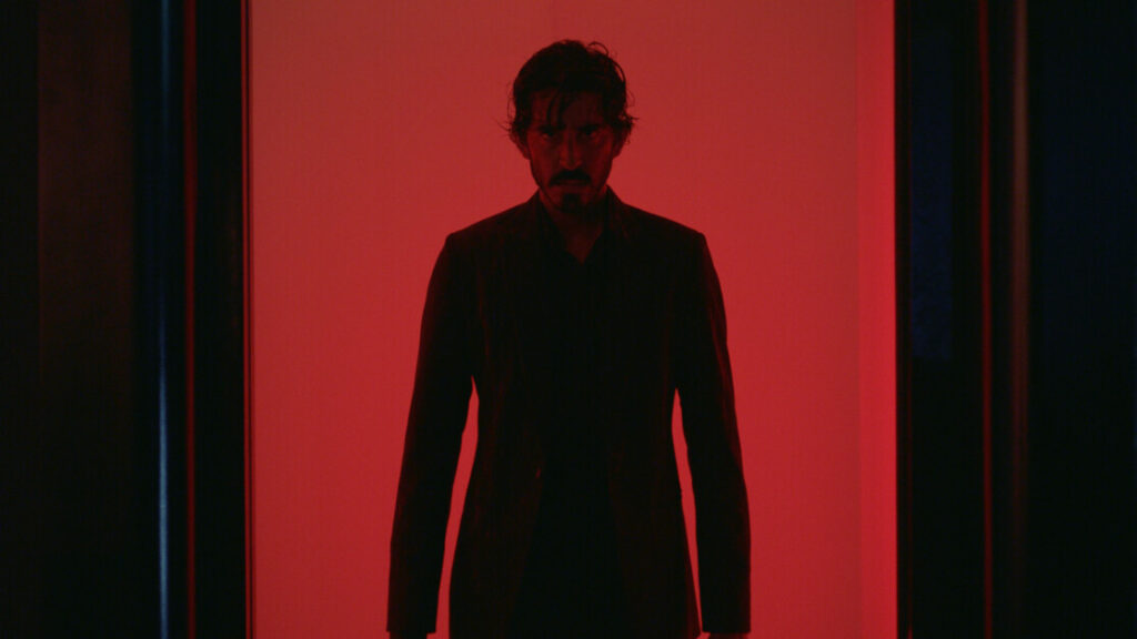 A man with dark hair and a beard stands in a dimly lit hallway bathed in red light, looking forward with a serious expression. He is wearing a dark suit.