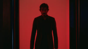 ‘Monkey Man’ 4K UHD Blu-Ray Review – A Brutal, Impressive Debut From Dev Patel A man with dark hair and a beard stands in a dimly lit hallway bathed in red light, looking forward with a serious expression. He is wearing a dark suit.