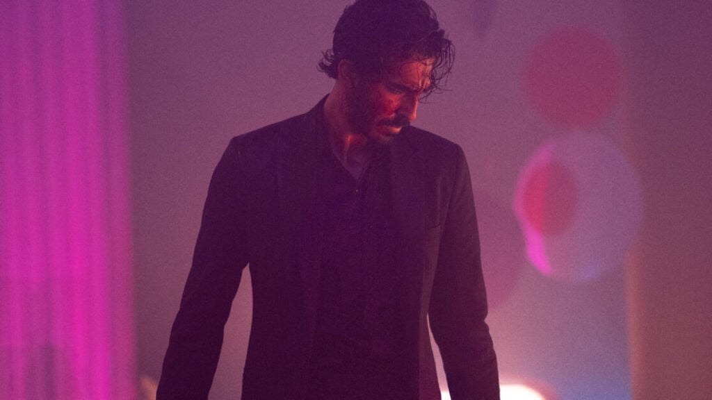 A person with wet, tousled hair stands in a dimly lit room wearing a dark suit, looking downwards. The background features blurred, colorful lights and pinkish hues.