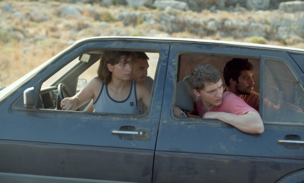 Four people are looking out of the open windows of an old, dusty car stopped in a rocky, desert-like area. The driver is holding the steering wheel with two passengers looking back.
