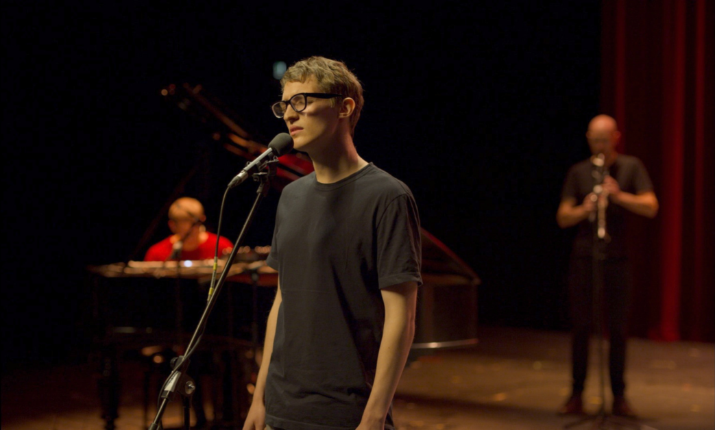 A person in a black shirt stands and sings into a microphone. A pianist and a clarinet player perform in the background on a dimly lit stage.