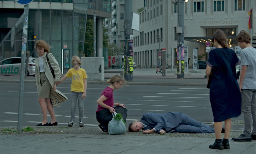 A man is lying on the ground with a small group of people gathered around him on a city street corner. One person is kneeling next to him, while others are standing and watching.