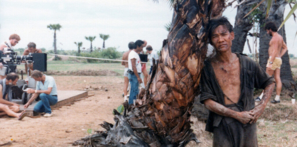 A man leans against a tree with a tired expression, while a film crew sets up equipment and interacts in the background. The setting appears to be a dry, rural area with sparse vegetation.