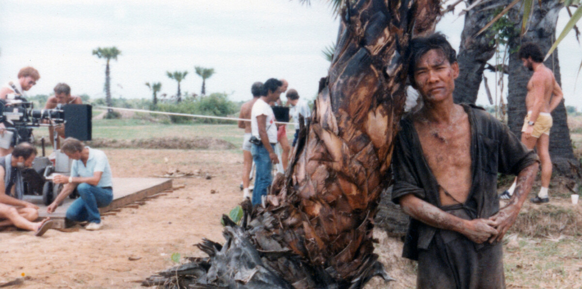 A man leans against a tree with a tired expression, while a film crew sets up equipment and interacts in the background. The setting appears to be a dry, rural area with sparse vegetation.