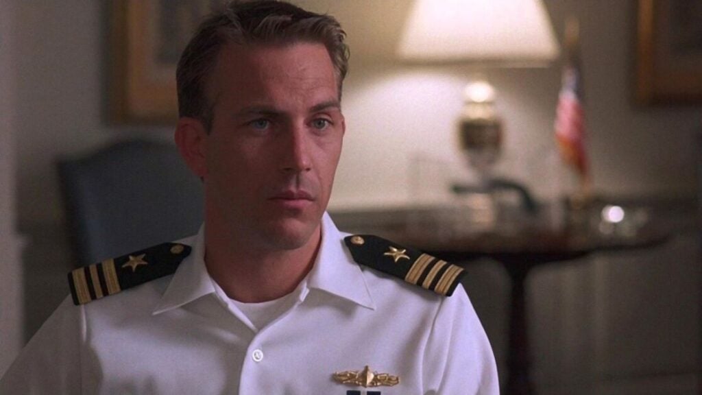 A man in a white naval uniform with two gold stars on the shoulder stands in a room with an American flag and a lamp in the background.