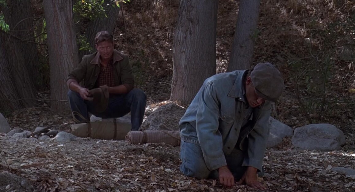 Two men are outdoors among trees. One is squatting and examining the ground while the other sits on a rock, holding a hat and a blanket roll. Fallen leaves cover the ground.