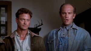 ‘Of Mice And Men’ (1992) Blu-Ray Review – A Respectful Adaptation Of The Classic Novel Two men, one with short curly hair in a brown jacket and the other bald in denim overalls and a blue shirt, stand side by side indoors with a deer antler mount visible in the background.
