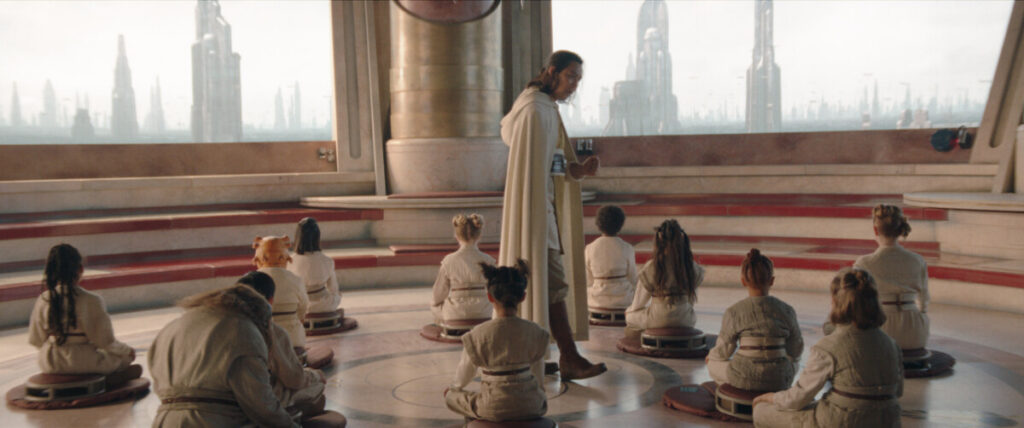 A teacher in a white robe instructs a group of children seated on the floor in a futuristic classroom with large windows revealing a cityscape.