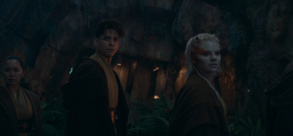 Three individuals wearing brown robes stand in a dark forest with a stone structure behind them, looking ahead with serious expressions.