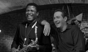 A man holding a saxophone stands beside another man; both are smiling and leaning on a brick wall in a black-and-white photograph.