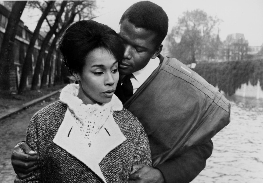 A man stands embracing a woman from behind along a waterfront, both dressed in mid-20th-century attire. The woman appears thoughtful, and trees line the background.