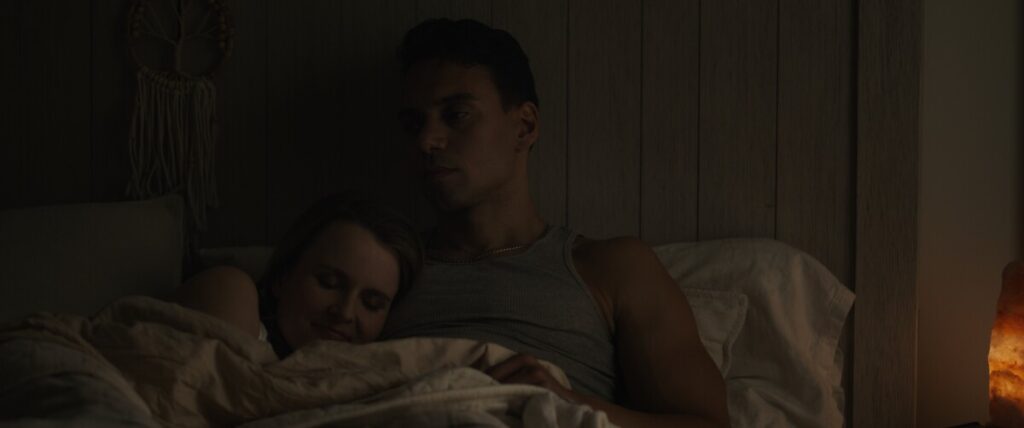 A couple sits close together in a dimly lit room, with the woman resting her head on the man's chest while lying in bed.